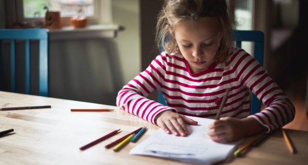A 7 year old left-handed girl is using colored pencils to homework. She is seated at a table near windows.